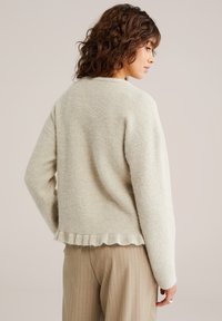 Pulover bej tricotat cu finisaj texturat și tiv ondulat. Guler rotund și umeri lăsați; purtat peste pantaloni bej cu dungi subțiri.