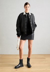 Veste bomber noire avec col blanc, jupe noire ajustée et bottines noires. Tenant un sac à main de couleur claire, debout sur un sol en bois.
