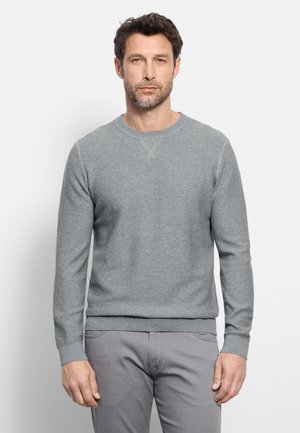 Mann mit kurzen braunen Haaren und Bart, trägt einen eng anliegenden hellgrauen gerippten Pullover und graue Hose, steht vor einem schlichten weißen Hintergrund.