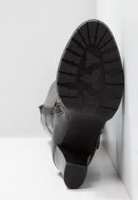 Botte en cuir noir avec semelle rainurée épaisse et fermeture éclair sur le côté, positionnée talon vers le haut contre un mur d'angle blanc.