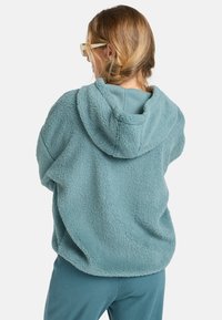 Een teal fleece hoodie met een gestructureerd oppervlak en een grote capuchon, gecombineerd met bijpassende geribbelde broek. De stof lijkt zacht en comfortabel.