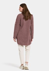 Bordeaux fleecetunika med en blød tekstur, lange ærmer og en høj-lav kant, helt båret med cremefarvede tapered bukser og hvide sneakers.