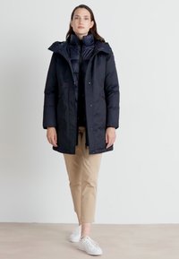 Lang, mørk marineblå pufferjakke med hette, featuring glidelås og ribbede mansjetter, lagt over en kviltet svart indre jakke. Brune bukser og hvite sneakers.