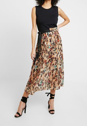 Kobieta w czarnym topie bez rękawów, plisowanej, średniej długości spódnicy w panterkę z czarnym paskiem oraz czarnych sandałach na wysokim obcasie z wiązaniami wokół kostki.