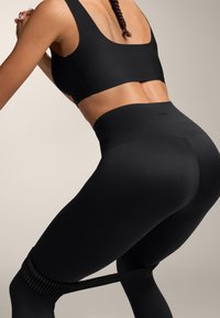 Nő fekete sportmelltartót és magas derekú leggingset visel, miközben ellenállás szalagot használ a combjai körül edzés közben.