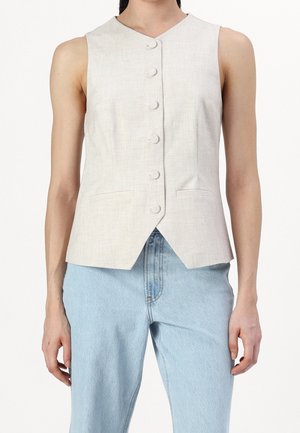 Beżowa bezrękawnik zapinany na guziki z ostrym dołem, noszony na jasnoniebieskich dżinsach na stojącej osobie.