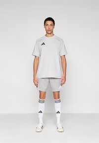 Ανοιχτό γκρι αθλητική φανέλα και σορτς με μαύρο λογότυπο Adidas. Λευκές κάλτσες μέχρι το γόνατο με μαύρες ρίγες. Μαύρα και λευκά ποδοσφαιρικά παπούτσια.