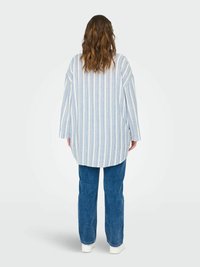 Chemise oversize rayée en bleu clair et blanc, en tissu doux, avec de larges manches et un ourlet arrondi. Associée à un jean bleu ample.
