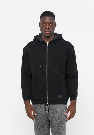 Black zip-up hoodie with a front pocket, drawstring hood, and ribbed cuffs. Thick fabric with smooth texture. Worn with grey patterned jeans.