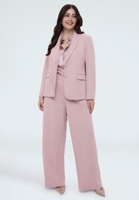 Lyserødt jakkesæt med en tætsiddende blazer og bukser med vidde, kombineret med en silkeagtig bluse. Har knappelementer og en glat tekstur.