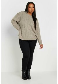 Suéter de punto beige con hombros caídos, combinado con jeans ajustados negros y botas de tobillo negras, con un fondo liso.