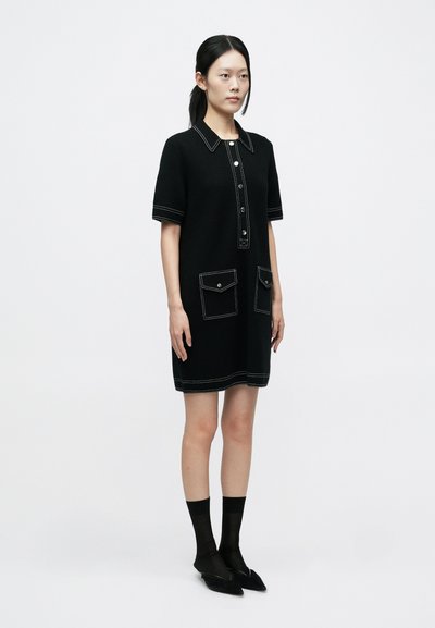 Fekete, rövid ujjú ruha gallérral, kontrasztos fehér varrással, két elülső zsebbel és gombos záródással. A sziluett egyenes.