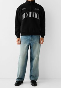 Svart fleece-tröja med halvdragkedja, med "BUSHWICK" i stora vita bokstäver. Kombinerad med ljusblå raka jeans och svarta skor.