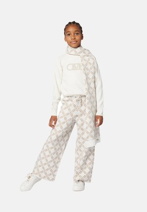 Maglione bianco a maglia con logo, abbinato a pantaloni crema con motivo geometrico e sneakers bianche. Sciarpa drappeggiata attorno al collo.