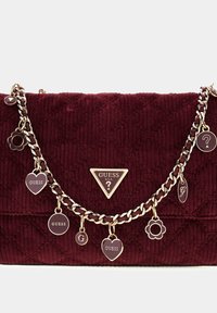 Burgundowa torebka z pikowanego materiału z złotym łańcuchem i zawieszkami, z motywami serc, kwiatów, kół oraz emblematami i metkami Guess.
