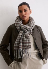 Écharpe à carreaux marron et bleu clair en mélange de laine, avec des bords frangés, portée sur un pull beige et sous une veste en cuir noire.