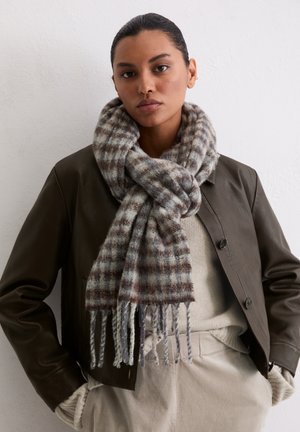 Écharpe à carreaux marron et bleu clair en mélange de laine, avec des bords frangés, portée sur un pull beige et sous une veste en cuir noire.