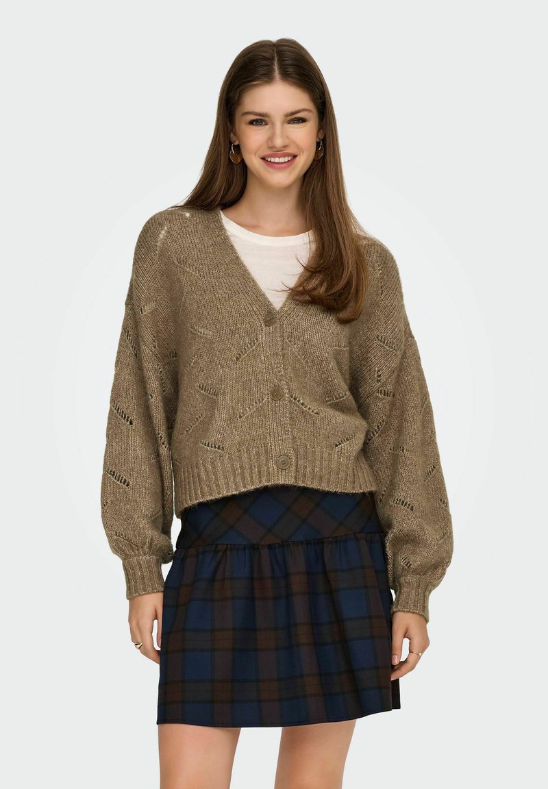 Beige strikket cardigan med v-udskæring, lange ballonærmer og et struktureret mønster, parret med en mørk ternet nederdel i blå og brune nuancer.