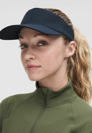 Frau mit blondem Haar im Pferdeschwanz, die einen schwarzen Visor und ein grünes, strukturiertes Zip-Up-Top trägt, schaut leicht seitlich mit neutralem Gesichtsausdruck.