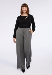 Eine Frau steht und lächelt, trägt ein schwarzes Langarmoberteil mit Ausschnitt und hochtaillierte graue weite Hose mit schwarzen spitzen Schuhen.