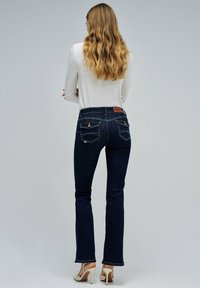Jean de campana en azul oscuro con un talle medio, que presenta dos bolsillos traseros con costuras, combinados con una blusa blanca de manga larga.