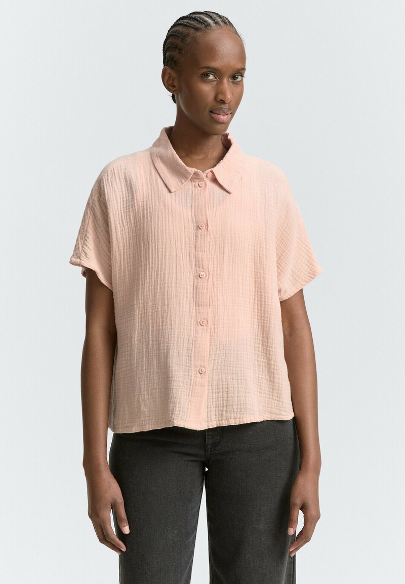 Camicia a maniche corte con bottoni in morbido tessuto color pesca, con una texture delicata. Presenta un colletto classico e una vestibilità comoda, abbinata a pantaloni scuri.