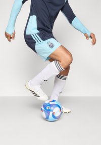 Ποδοσφαιρική στολή σε ναυτικό και ανοιχτό μπλε χρώμα με μακριά μανίκια, συνδυασμένα σορτς και λευκές κάλτσες. Παίκτης χειρίζεται την μπάλα που διαθέτει γεωμετρικά σχέδια.