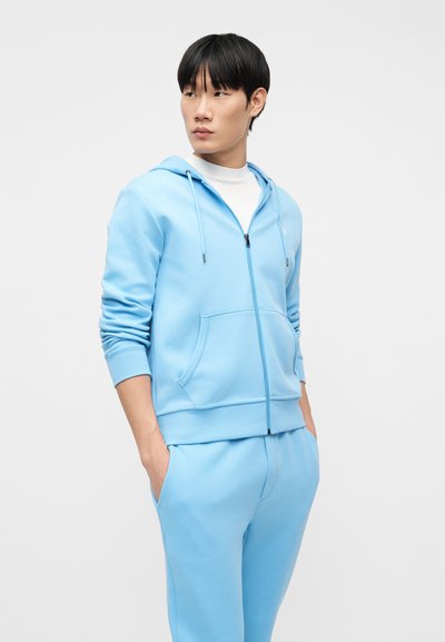 Νεαρός άνδρας που φοράει γαλάζιο φούτερ με φερμουάρ και ταιριαστό παντελόνι με τα χέρια στις τσέπες, κοιτάζοντας προς το πλάι μπροστά σε απλό φόντο.