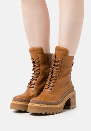Bottines marron en cuir à lacets avec talons épais et semelles rainurées, portées sur des jambes nues sur fond blanc.