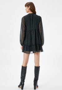 Schwarzes, gepunktetes, gestuftes Minikleid mit transparenten Ärmeln, Rüsche am Saum und Rückenschluss, kombiniert mit knielangen schwarzen Lederstiefeln.