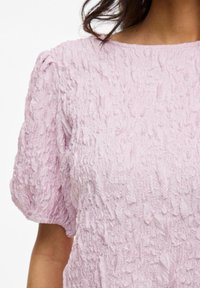 Blusa leggera di un rosa pastello con maniche a palloncino e design in tessuto arricciato. Presenta una morbida texture stropicciata e un colletto rotondo.