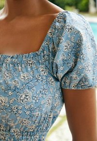 Blusa floral azul con mangas abullonadas, que presenta un escote cuadrado y detalles fruncidos. El patrón incluye flores blancas y hojas.