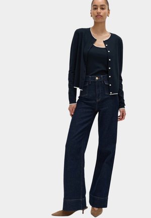 Vrouw die een donkerblauw gebreid vest met knopen draagt over een bijpassende tanktop, wijde jeans met hoge taille en bruine puntige hakken, staand tegen een witte achtergrond.