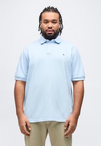 Άνδρας με πλεγμένα μαλλιά και γενειάδα, που φοράει ανοιχτό μπλε πόλο πουκάμισο και μπεζ παντελόνι, στέκεται μπροστά σε λευκό φόντο.