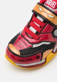 Rote und schwarze Sneaker mit glänzender Oberfläche, Mesh-Einsätzen und gelbem Außensohlenrand. Akzentuiert mit silbernen Details und einem Marvel-Logo.