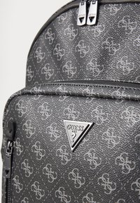 Gray faux leather backpack with patterned logo, featuring a prominent triangular Guess logo and silver zippers. Textured surface detail.