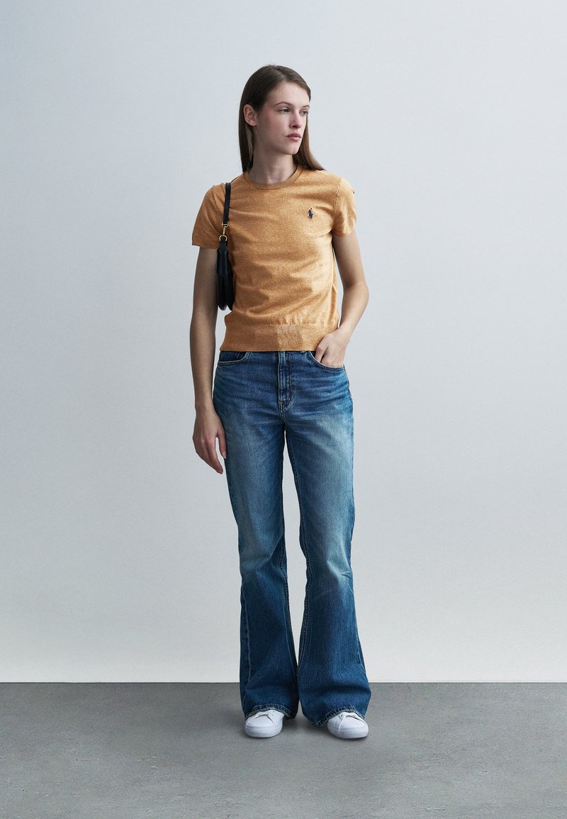 Haut orange à manches courtes en tricot avec une taille ajustée, associé à un jean bleu délavé évasé et des baskets blanches, le tout complété par un sac à main noir.