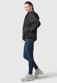 Black lightweight jacket with a hood, zip closure, and elastic hem, paired with blue skinny jeans and neutral-toned sneakers.
