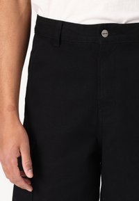 Pantaloni negri din bumbac cu închidere cu nasturi și buzunare laterale; țesătură texturată cu detalii de cusături vizibile. Mână odihnindu-se aproape de talie.