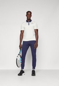 Bijela polo majica s tamnoplavim ovratnikom i detaljima, uparena s tamnoplave jogger hlačama. Model drži tenisku rekete i nosi crnu sportsku obuću.
