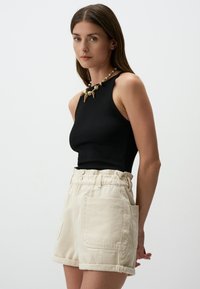 Top aderente nero con scollatura alta e collana decorativa beige, abbinato a pantaloni corti beige chiaro a vita alta con tasche laterali.
