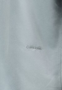 Ανοιχτό γκρι ύφασμα με διακριτικό ανάγλυφο λογότυπο Calvin Klein στο κέντρο, που δείχνει λεπτομέρειες υφής και ραφής στην δεξιά άκρη.
