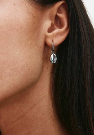 Srebrna kolczyk-koło z polerowanym zawieszką z muszli kauri noszony na lewym uchu osoby z widocznymi ciemnymi włosami.