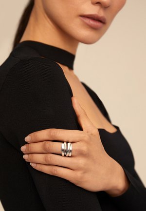 Mujer con un top negro de cuello choker, sosteniendo su brazo y mostrando un anillo ancho de plata de triple banda en su dedo medio.