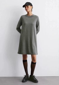 Donna che indossa un abito lungo a maglia grigio con maniche lunghe, cappello nero, calze nere trasparenti fin sopra il ginocchio e scarpe con piattaforma nere, in piedi contro un muro bianco.
