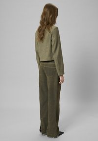 Oliivroheline lühike jakk ja laiade jalgadega teksapüksid tekstureeritud denimist, millel on kulunud servad ja diskreetsed tagakoti aksessuaarid.