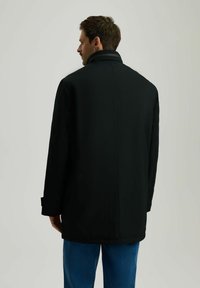 Czarny płaszcz z wysokim kołnierzem, prostym krojem i bocznymi panelami, w połączeniu z niebieskimi dżinsami. Gładka tekstura; minimalistyczny design bez widocznych elementów metalowych.