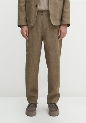 Person iført brune strukturerede jakkesætsbukser, matchende blazer, beige sweater og brune ruskindssnøresko stående på hvidt gulv.