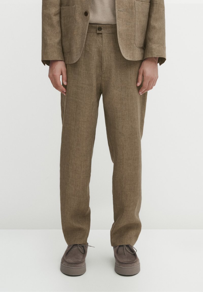 Personne portant un pantalon de costume marron texturé, un blazer assorti, un pull beige et des chaussures en daim marron à lacets, debout sur un sol blanc.