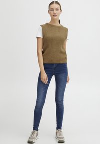 Brun ribbestrikket ermeløs topp over en hvit t-skjorte, sammen med trange blå jeans og lyse beige sneakers med grå detaljer.